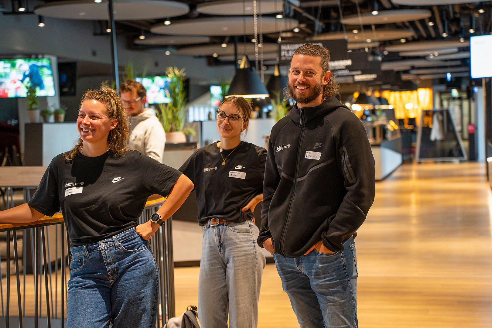 Drei Personen der beruf leben akademie im Europapark-Stadion beim Fachtag, lächeln, tragen einheitliches schwarzes T-Shirt der Sport-Quartiere Freiburg.